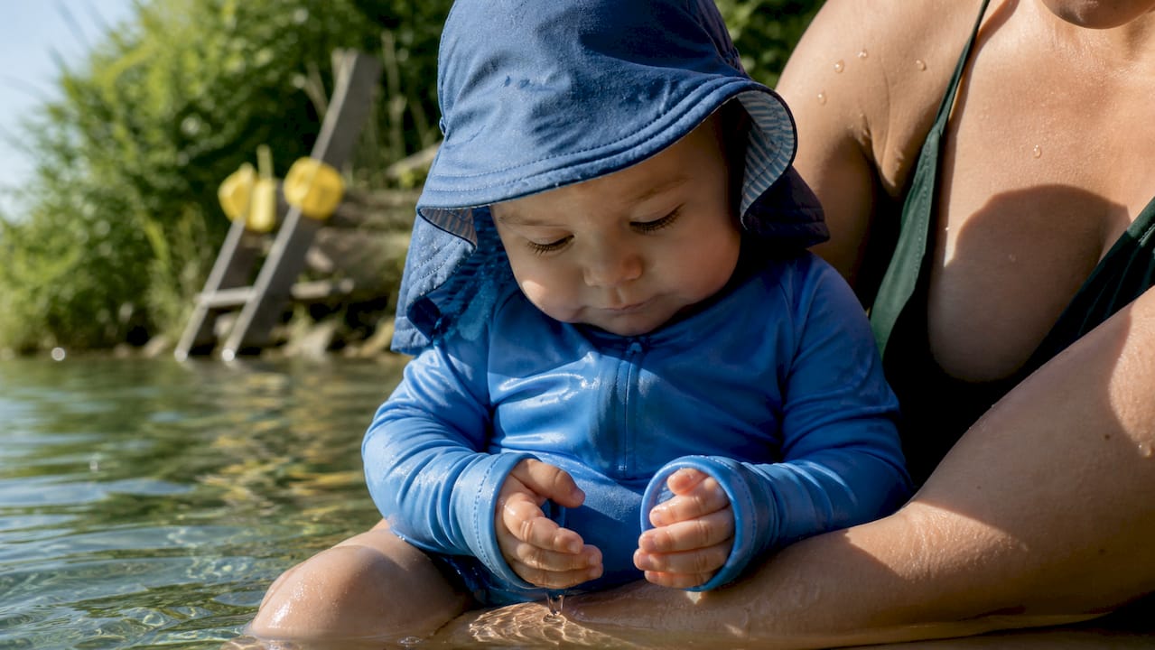 Ein Baby wird am Teich gehalten und trägt dabei ein UV-Schutzshirt sowie einen Sonnenhut.