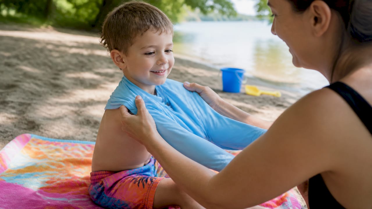 Am Strand zieht ein Kind gerade ein spezielles Sonnenschutzshirt mit UPF an, damit es beim Spielen optimal vor der Sonne geschützt ist.